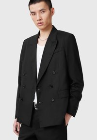 Blazer nero doppiopetto con rever a punta, due tasche frontali e sei bottoni neri. Indossato sopra una maglietta bianca. Tessuto liscio.