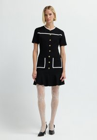 Abito nero a maniche corte con fondo svasato, dettagli bianchi, bottoni dorati e tasche frontali. La modella indossa collant a fantasia e tacchi neri.