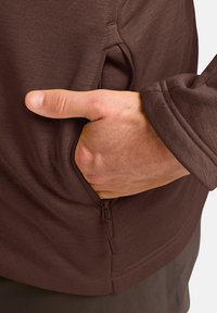 Bruine lange mouw kledingstuk met een zijzak en ritssluiting. Zachte stofstructuur en een getailleerd ontwerp met een subtiele glans.