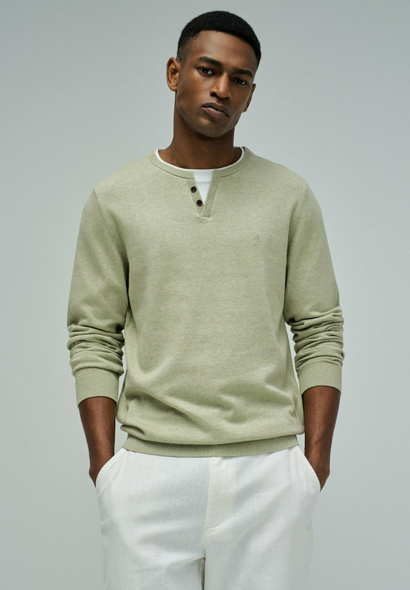 Junger Mann, der einen hellgrünen Strickpullover über einem weißen Hemd trägt, mit den Händen in den weißen Hosentaschen vor einem schlichten grauen Hintergrund.