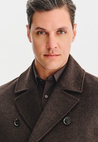 Brązowy wełniany płaszcz z dużymi wyłogami i czarnymi guzikami, noszony na ciemnej koszuli. Posiada strukturalny materiał z dopasowanym krojem.