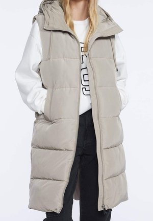 Vrouw draagt een beige gewatteerde mouwloze capuchonvest over een witte sweatshirt en zwarte broek, met haar handen in de zakken van het vest.