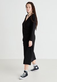 Robe longue noire à manches longues, avec un décolleté en V profond et un design drapé, associée à des baskets montantes noires et des chaussettes grises. Tissu simple et lisse.