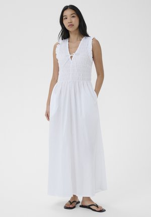 Nő ujjatlan fehér maxi ruhában, melynek felső része ráncolt, V-nyakú és zsebes, fekete papuccsal párosítva, egy egyszerű hátteret nézve áll.