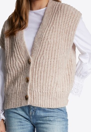Frau trägt beige grob gestrickte, geknöpfte Weste über weißer Bluse mit langen Ärmeln und Lochspitzenmanschetten sowie blaue Jeans, gezeigt vom Hals bis zur Hüfte.