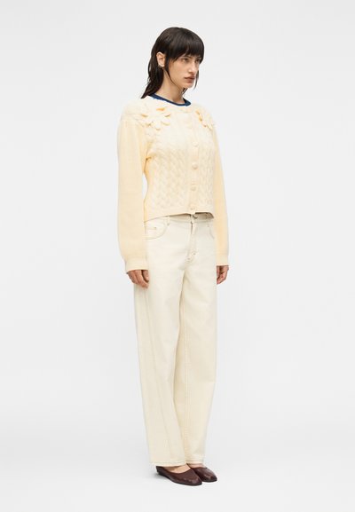 Γυναίκα με σκούρα μαλλιά που φοράει κρέμ ζακέτα πλεχτή με ανάγλυφα λουλούδια, λευκό παντελόνι με φαρδύ πόδι και σκούρα καφέ επίπεδα παπούτσια, που στέκεται πλάγια.