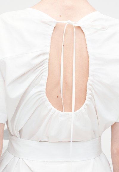 Πίσω μέρος προσώπου που φοράει λευκό φόρεμα με πιέτες γύρω από ένα οβάλ άνοιγμα δεμένο στο πάνω μέρος με λεπτά λευκά κορδόνια.