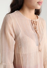 Transparente Bluse in Blush mit rundem Ausschnitt und Bindedetail, ausgestattet mit einem strukturierten Muster und langen Ärmeln. Leichter Stoff mit dezenten Streifen.