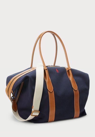 Saco de duffel de lona azul-marinho com detalhes e pegas em couro castanho, apresentando uma alça ajustável em creme e um pequeno detalhe de logotipo vermelho. Fecho com zíper.