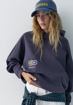 Mladi posameznik z temno kapuco z napisom "Creative for Life", modro kapo Michigan in srajco s črtami, zavezano okoli pasu.