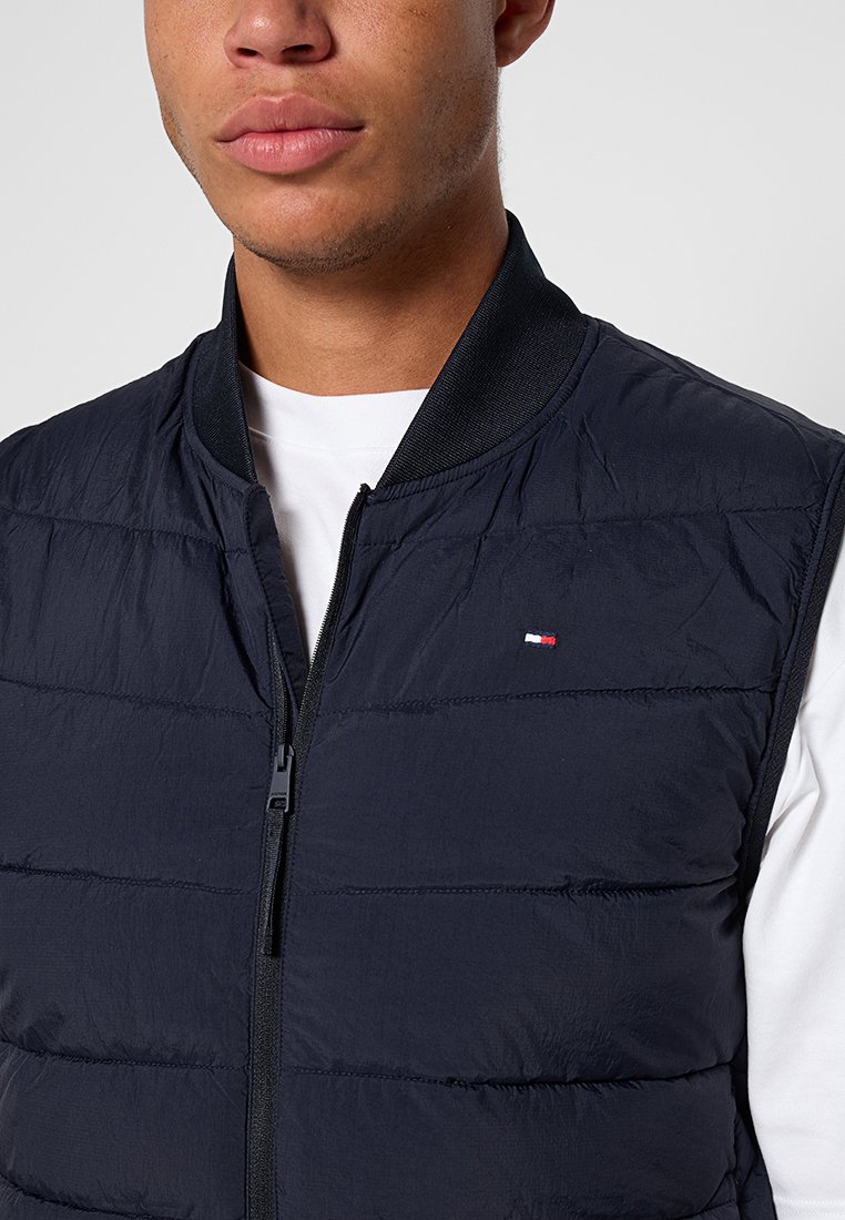 Chaleco acolchado azul marino con cierre de cremallera, cuello redondo y un pequeño logo. Presenta una textura suave y costuras horizontales en el cuerpo.