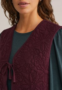 Gilet bordeaux imbottito con motivo floreale, dotato di chiusura a lacci nella parte anteriore, indossato sopra una maglietta a maniche lunghe di color teal scuro.