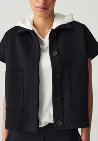 Schwarze kurzärmelige Jacke mit strukturiertem Design, vorderen Knöpfen, zwei großen Taschen und einer Kapuze, die über einem weißen gerippten Oberteil getragen wird.
