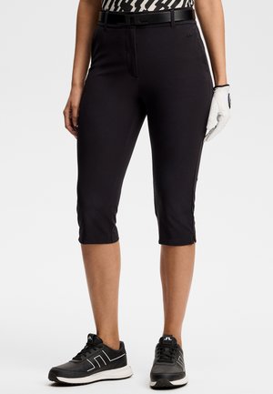 Vrouw draagt zwarte cropped broek, zwarte riem, patroon top, zwarte sneakers en een witte golfhandschoen aan de rechterhand, staand tegen een witte achtergrond.