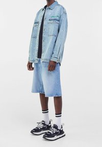 Ljust blå denimjacka med två framfickor, med grön broderi och färgsprut. Ihop med matchande blå denimshorts och svarta sneakers.