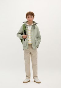 Lichtgroene waterdichte jas met een capuchon, wit en geel gestreept shirt, beige cargobroek en witte schoenen met een gumrubberen zool.