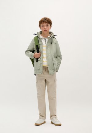 Lichtgroene waterdichte jas met een capuchon, wit en geel gestreept shirt, beige cargobroek en witte schoenen met een gumrubberen zool.