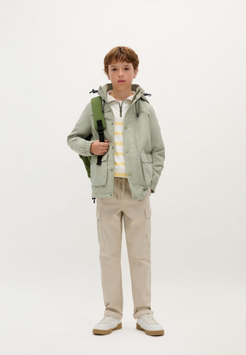 Lichtgroene waterdichte jas met een capuchon, wit en geel gestreept shirt, beige cargobroek en witte schoenen met een gumrubberen zool.
