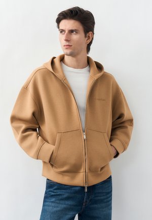 Mladý muž v béžovej mikine na zips a bielom tričku, stojaci s rukami v vreckách, hľadí do strany na jednoduchom pozadí.