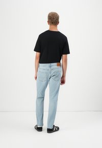 Mies, jolla on kiharaiset hiukset, pukeutunut mustaan t-paitaan, vaaleansinisiin farkkuihin ja mustiin tennareihin, seisoo valkoista seinää vasten.