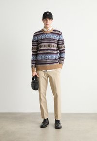 Pullover mit mehrfarbigen geometrischen Mustern, beigen Akzenten, kombiniert mit beigen Hosen und schwarzen Stiefeln. Hält eine kleine schwarze Tasche.