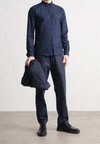 Mörkblå skjorta med knappar och krage, tillsammans med matchande byxor och en jacka i handen, allt i ett lätt tyg. Svarta skor.