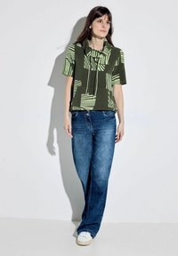 Maglietta verde a fantasia con maniche corte e collo a cowl, abbinata a jeans in denim blu e scarpe da ginnastica bianche. Tessuto liscio e vestibilità casual.