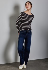 Gestreifter, schwarz-weißer, gerippter Pullover kombiniert mit dunkelblauen, weiten Jeans und weißen Sneakern, vor einem neutralen grauen Hintergrund.