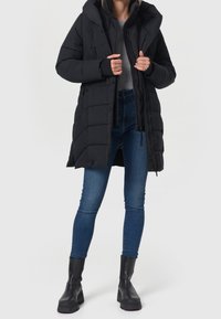 Sort pufferjakke med hætte, der har quiltet design, frontlynlås og sidelommer. Båret med blå jeans og sorte chunky støvler.