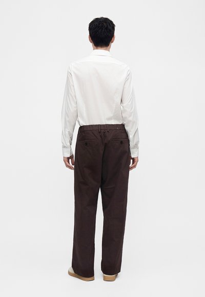 Homme de dos portant une chemise blanche à manches longues, un pantalon ample marron foncé et des chaussures beiges sur un fond clair uni.