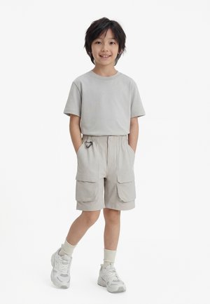 Boy with black hair wearing light gray t-shirt, beige cargo shorts with carabiner, cream socks, and white sneakers, smiling with hands in pockets.