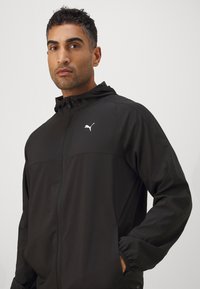 Schwarze Kapuzenjacke aus glattem Stoff mit einem Reißverschluss an der Vorderseite und einem weißen Puma-Logo auf der Brust. Elastische Bündchen und seitliche Taschen sind enthalten.