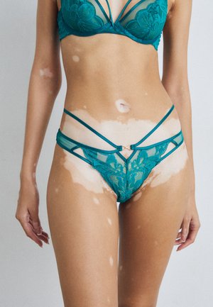 Teal lace bra and matching thong featuring floral patterns, sheer material, and multiple strap accents. Golden hardware detail at the waist.