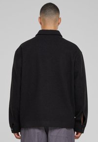Černá, oversize bunda s límcem, dlouhými rukávy a bočními rozparky. Má hladkou texturu a knoflíkové manžety.