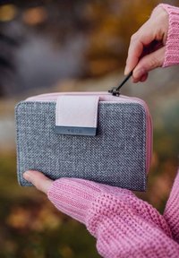 Gray textured fabric wallet with a light pink zipper and strap. Features a black metal logo plate. Hand holding it with a pink knitted sweater sleeve.