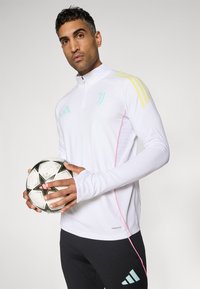 Witte sporttop met lange mouwen en ritskraag, met gele en turquoise accenten, gecombineerd met zwarte broeken, terwijl hij een voetbal vasthoudt.