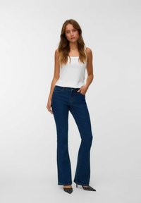 Kobieta stoi w białej bezrękawnikowej bluzce, ciemnoniebieskich, wysokich, rozszerzających się dżinsach i czarnych, spiczastych butach z białymi kropkami.