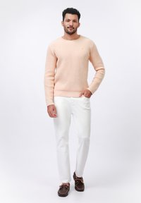 Light peach knitted sweater with round neckline, textured pattern, and small yellow accent logo. Paired with white pants and brown boat shoes.