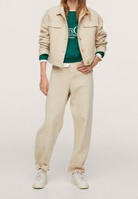 Veste en denim beige avec deux poches à rabat, associée à un sweat-shirt vert superposé à une chemise blanche et un pantalon large beige, complétée par des baskets blanches.