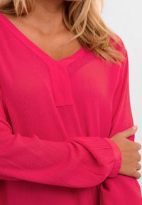 Blouse fuchsia avec un col en V et une texture côtelée. Manches longues réunies aux poignets, présentant un détail subtil de patte de boutonnage à l'avant.
