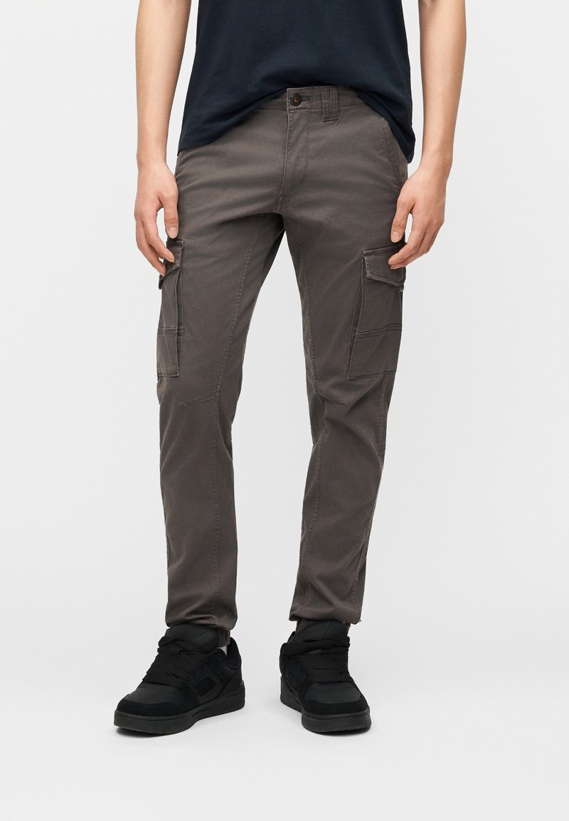 Personne portant un pantalon cargo gris foncé avec des poches latérales et des baskets noires, debout devant un fond clair uni.