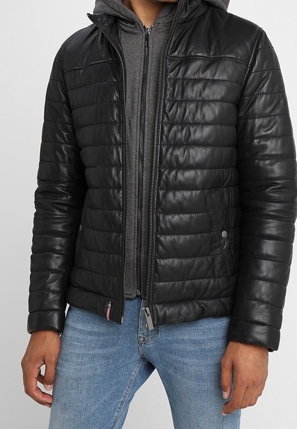 Man i svart quiltad läderjacka över grå zip-up hoodie och ljusblå jeans, med händerna avslappnat vid sidorna.