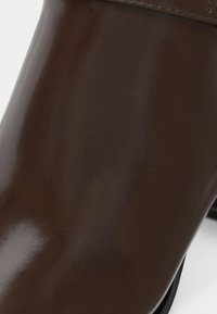 Botín de cuero marrón con un acabado brillante, que presenta una puntera redondeada y un diseño de costuras mínimas. Suela negra visible en la parte inferior.