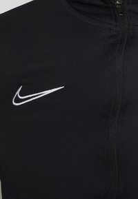 Zwarte sportjack van gestructureerde stof met een volledige ritssluiting aan de voorkant en een wit geborduurd Nike-swoosh op de linkerborst.
