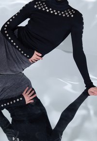 Schwarzer Rollkragenpullover mit metallicen Ösenakzenten an den Schultern, kombiniert mit einem dunklen, strukturierten Rock; auf einer glänzenden Oberfläche reflektiert.