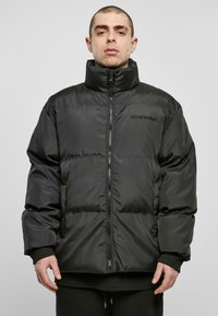 Jachetă puffer neagră cu guler înalt, închidere cu fermoar și siluetă elongated. Prezintă secțiuni matlasate și branding minimal pe piept.