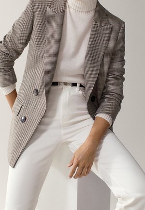 Woman in white turtleneck, high-waisted white pants, and checked double-breasted blazer with sleeves pushed up, posing against a plain background.