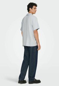 Kortærmet, lyseblå skjorte med krave og struktureret mønster, parret med mørkeblå vide jeans og sorte sko.