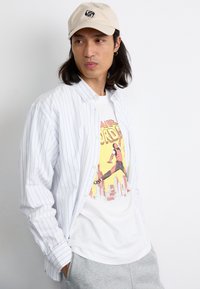Hvid skjorte med blå striber, over en grafisk hvid t-shirt. Lys beige kasket, grå sweatpants, og langt mørkt hår.