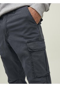 Personne portant un pantalon cargo gris foncé avec une poche à rabat, main reposant dans la poche avant, et manche de sweat-shirt gris clair visible.
